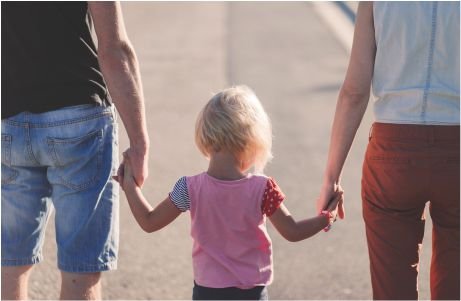 A child in a pink shirt holds hands with two adults.