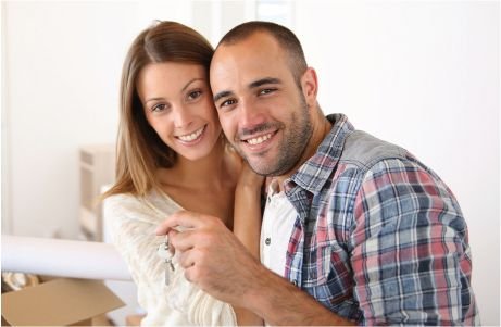 Smiling couple stand in a bright room, holding keys.