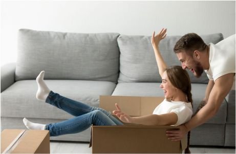 A man playfully pushes a smiling woman sitting in a cardboard box.