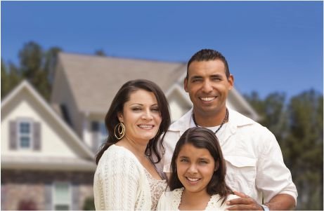 A smiling family of three stands together in front of a suburban house.